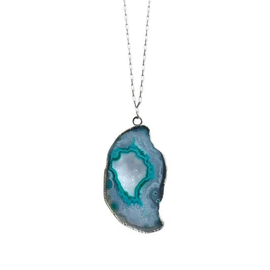 A silver necklace featuring a polished turquoise geode pendant with vibrant teal  banded patterns and white crystallized quartz center on a sterling silver paperclip chain
